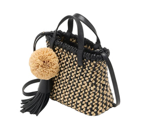 The Ulla Johnson Raffia Mini Crossbody is a small hand-woven tote with a black and beige checker pattern, black handles, and a black leather tassel accented by a large beige pom-pom.