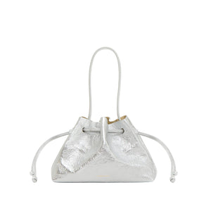 The Mansur Gavriel Drawstring Crinkle Pouchette is a metallic silver compact handbag with two side pull cords and a thin shoulder strap, crafted from premium leather and shown against a white background.