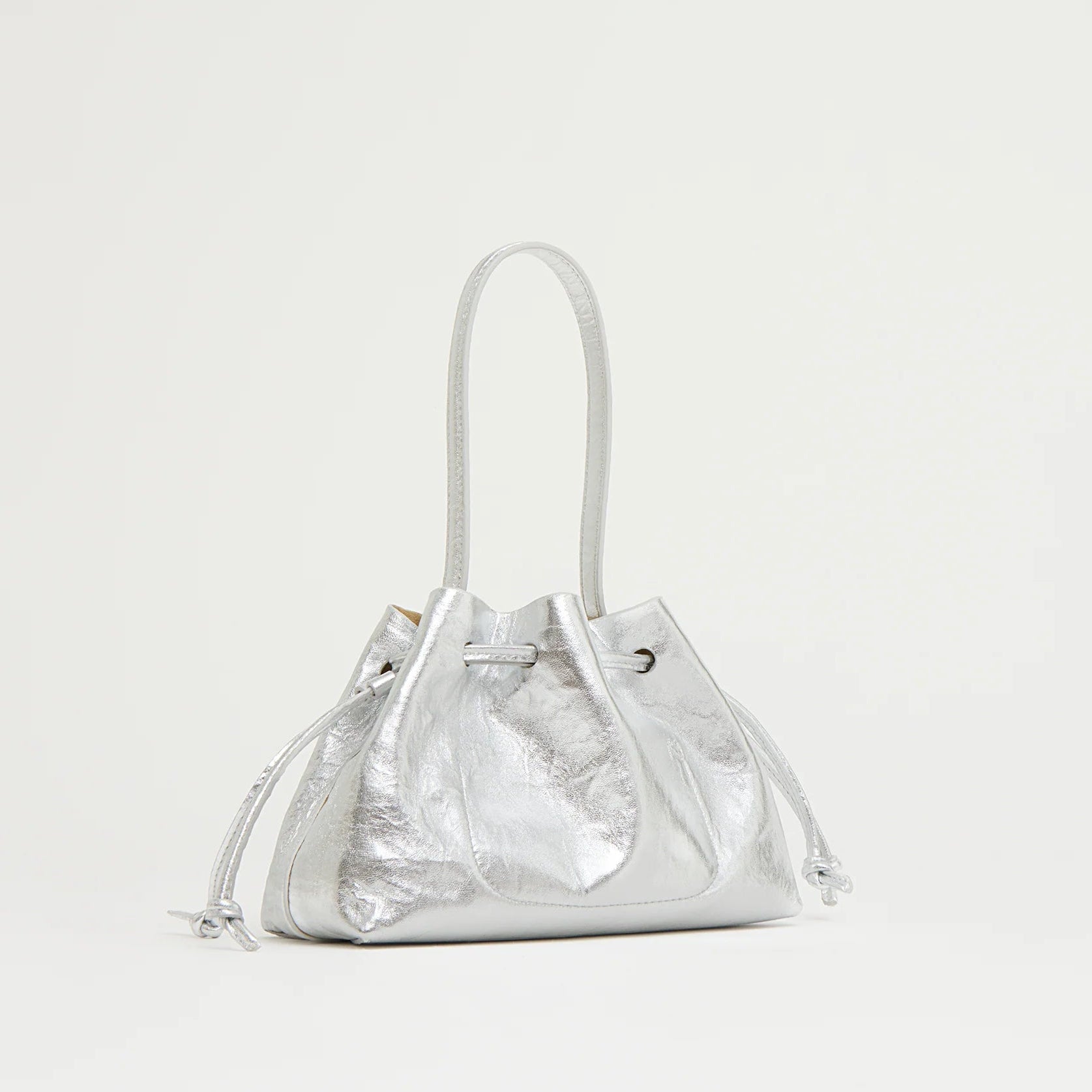 The Mansur Gavriel Drawstring Crinkle Pouchette is a small metallic silver Italian leather handbag with two handles and a gathered, textured design, shown against a plain light background.