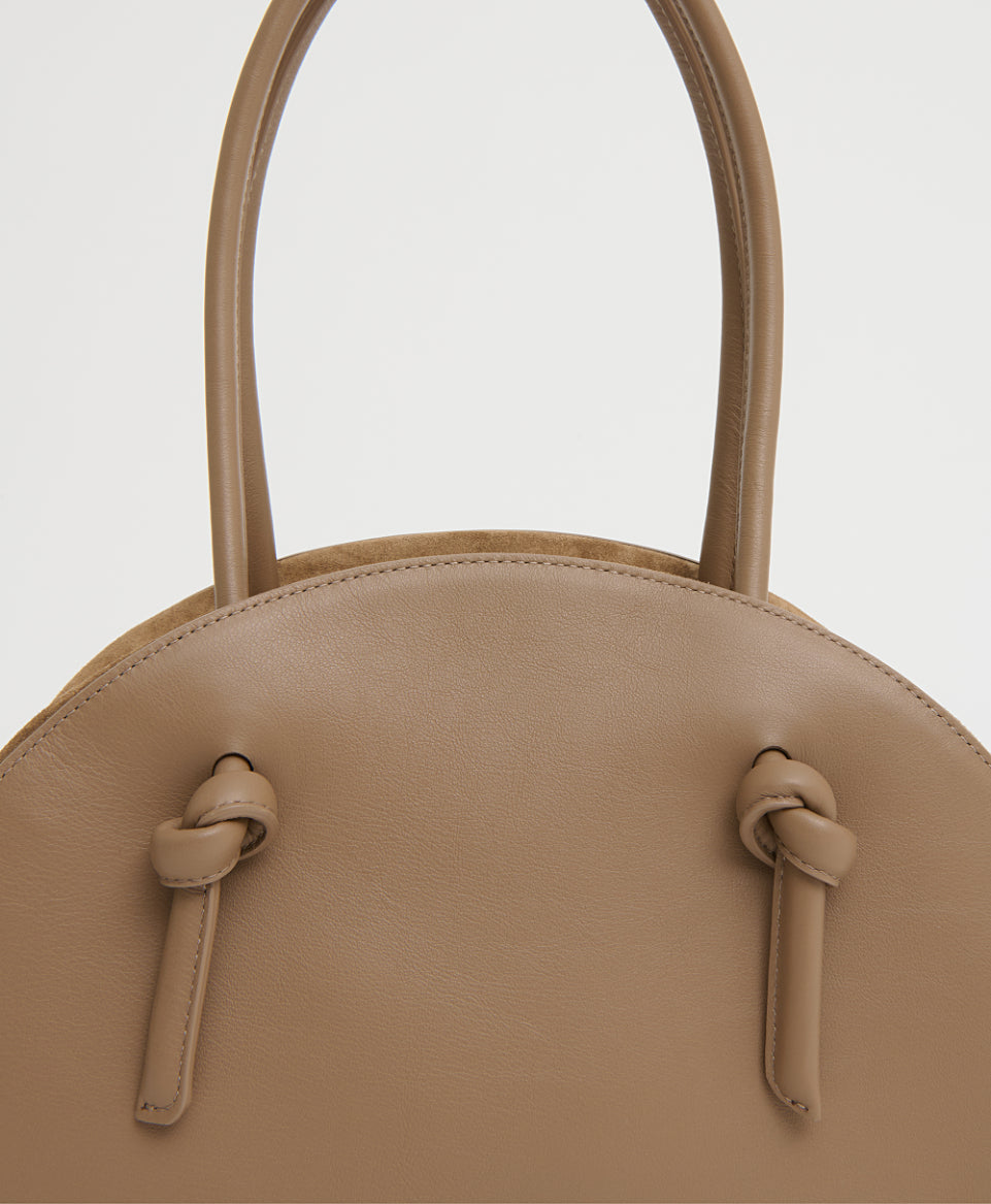 A close-up view of the Mansur Gavriel Fortuna Bowling Bag, a tan Italian leather top-handle bag featuring two handles and knotted strap details, shown against a plain background.