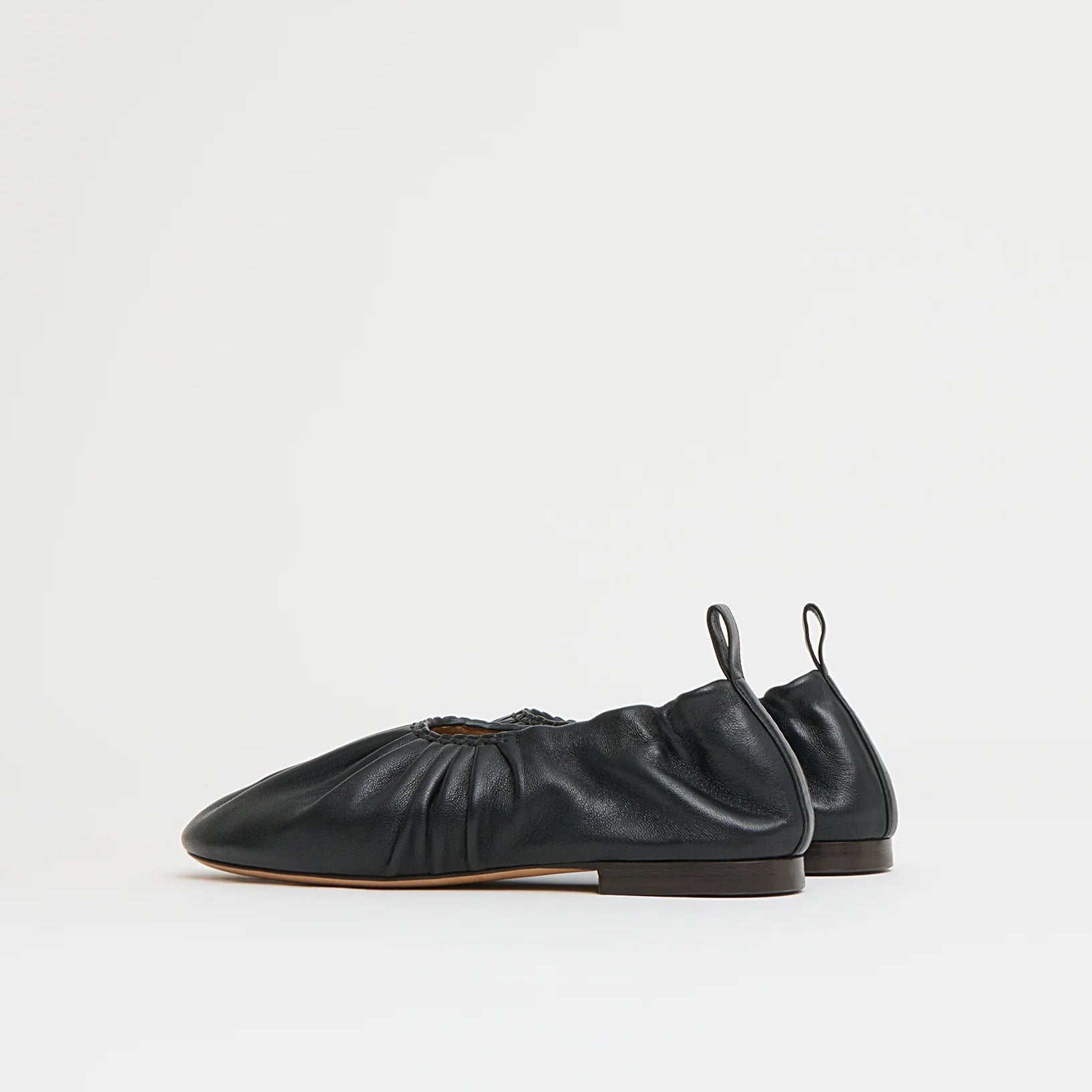 The Mansur Gavriel Ruched Ballerina, crafted from black Italian leather with gathered detailing, pull tabs at the heel, and flat soles, is shown against a plain white background.