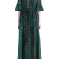 A model stands against a white background wearing the Altuzarra Thessaly Dress, a floor-length, dark green piece featuring short sleeves, a tie waist, and a subtle patterned design.