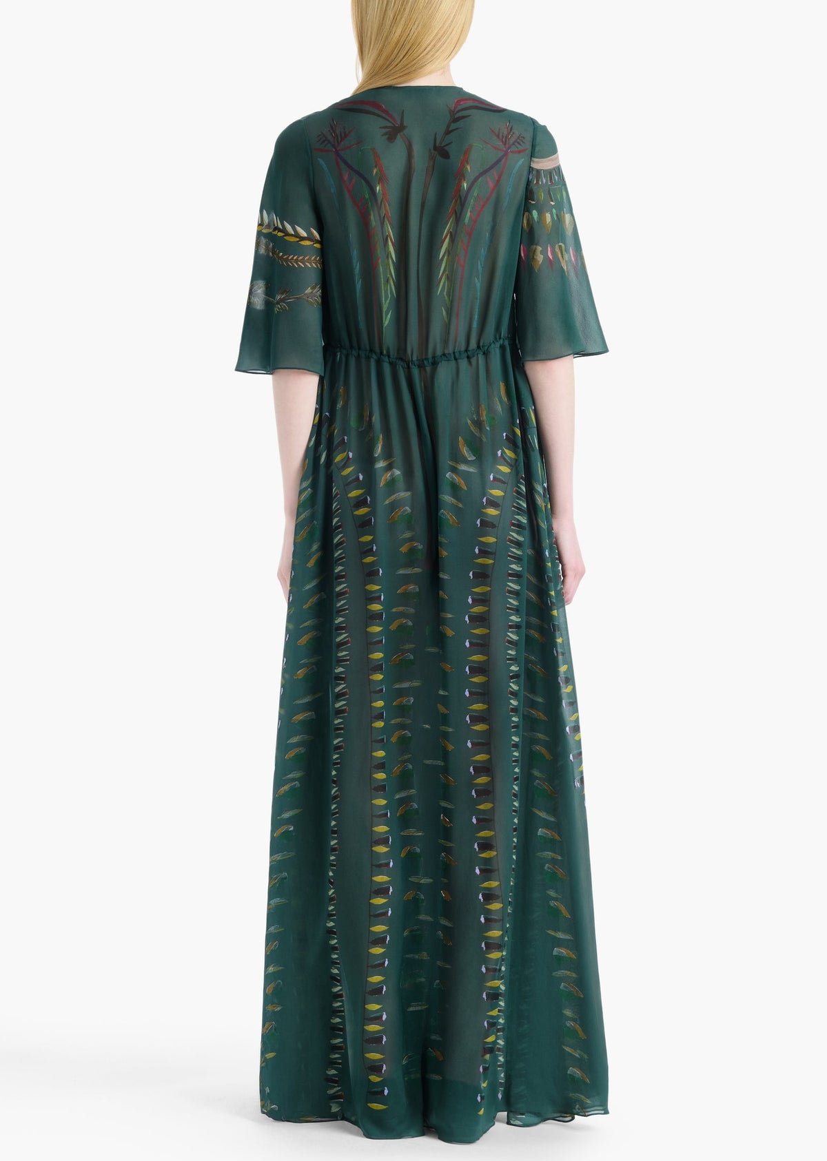A person is seen from the back against a plain white background, wearing the Thessaly Dress by Altuzarra—a long, sheer dark green silk dress featuring botanical patterns.