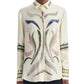 A person wearing the Altuzarra Chika Top—a cream-colored silk blouse featuring abstract multicolored patterns and embroidery on the front, collar, and sleeves, with a relaxed silhouette and a concealed button closure.
