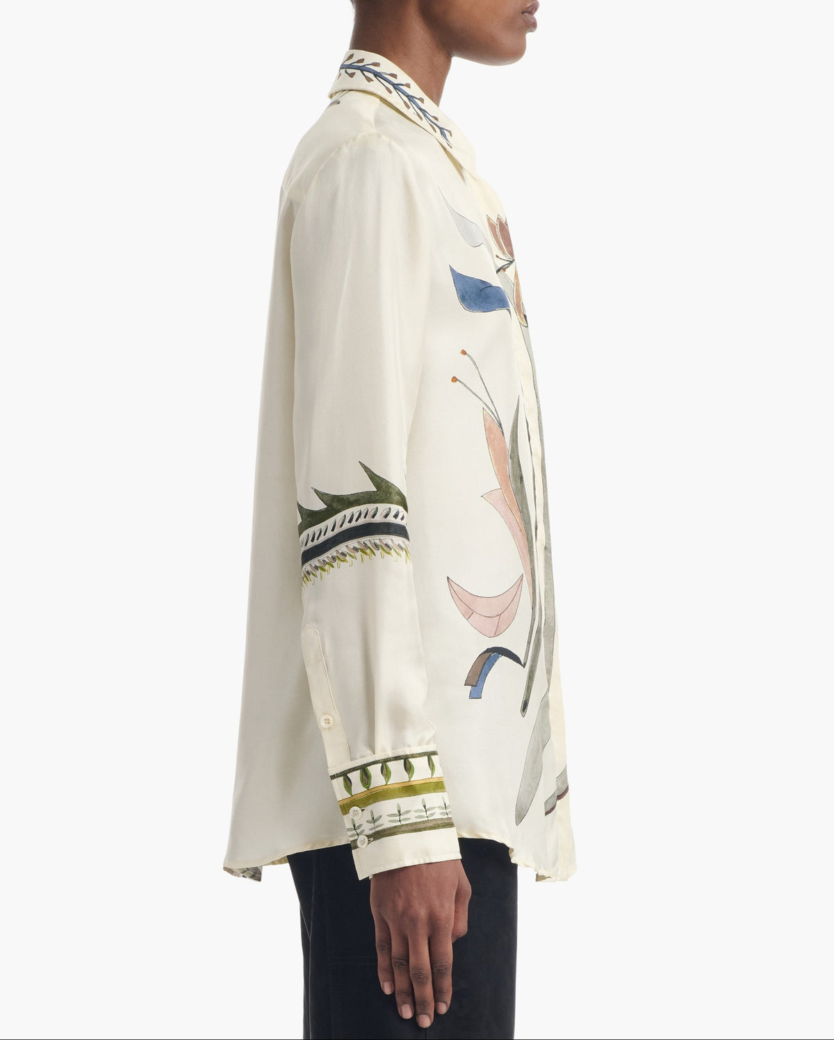 A person wearing the Altuzarra Chika Top, a relaxed long-sleeve cream silk blouse with colorful embroidered designs on the sleeves, front, and cuffs, is shown from the side against a white background.