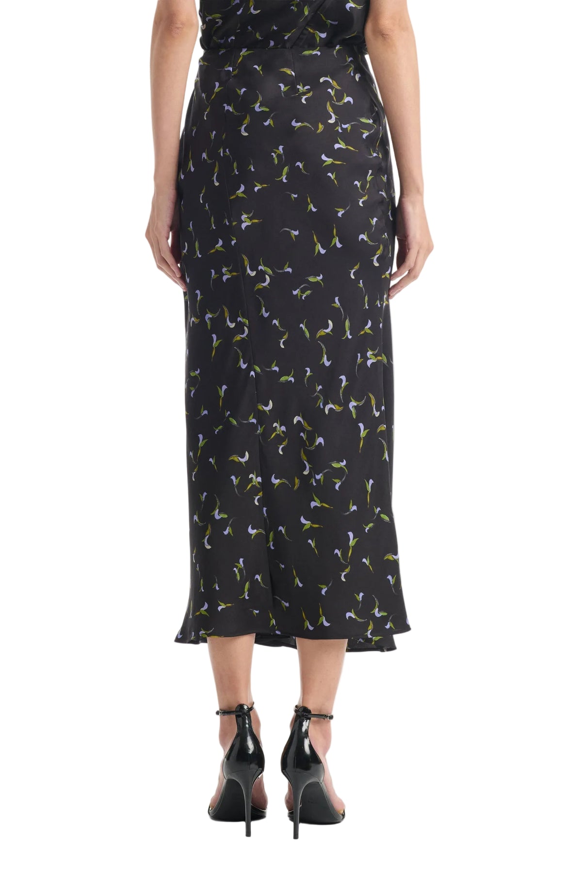 A person is shown from the back wearing the Altuzarra Casey Skirt, a black midi skirt with a small floral print and streamlined silhouette, paired with black high-heeled sandals.
