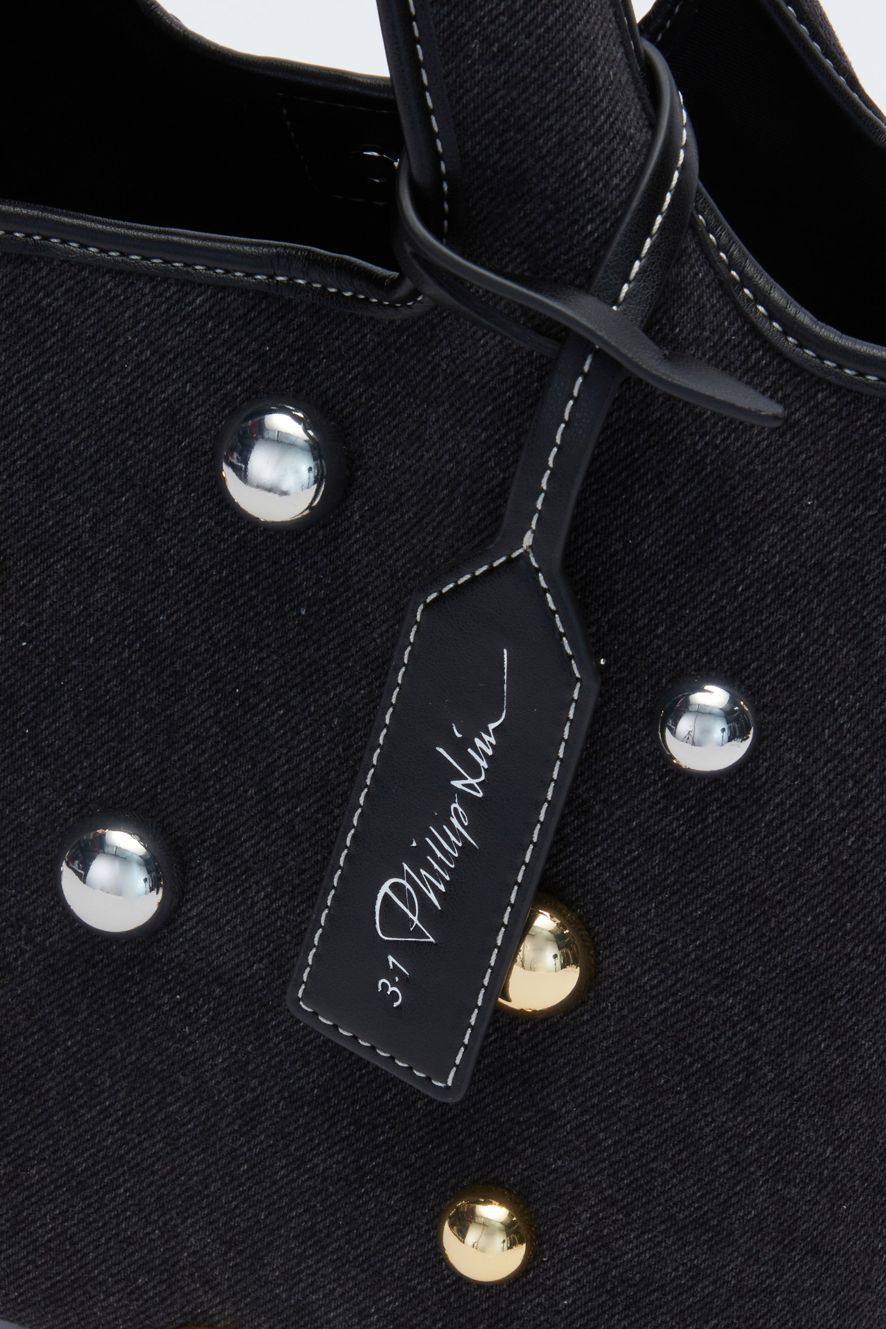 Close-up of a Washed Denim Mini Market Tote by 3.1 Phillip Lim, featuring silver and gold dome studs and a black tag with white branding.