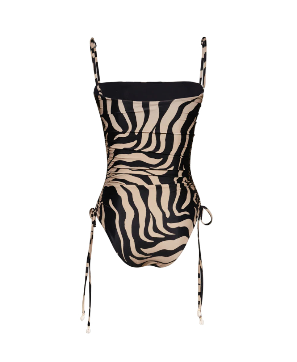 The Palms Tarangire One Piece by Johanna Ortiz is a beige and black zebra print swimsuit with a body-core silhouette, adjustable spaghetti straps, and ruching with side ties.