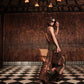 A woman wearing the Johanna Ortiz Encanto De Isla Ankle Skirt, sunglasses, and boots stands on a black-and-white checkered floor, holding a brown handbag in a warmly lit wooden room.