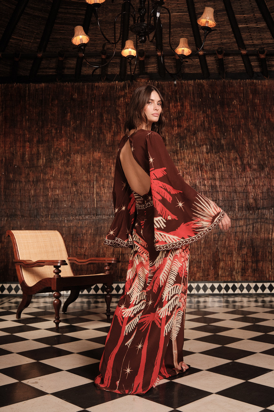 A woman wears the Johanna Ortiz Leyenda Del Tiempo Maxi Dress, featuring red and white leaf patterns and long sleeves, while standing on a black-and-white checkered floor with a wooden backdrop and chandelier.