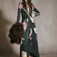 A model wears a dark green botanical Johanna Ortiz Reinvention Oasis Ankle Dress with a pink and cream floral print, plunging neckline, and knotted waist, paired with black boots and a brown fur stole.