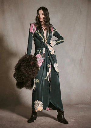 A model wears a dark green botanical Johanna Ortiz Reinvention Oasis Ankle Dress with a pink and cream floral print, plunging neckline, and knotted waist, paired with black boots and a brown fur stole.