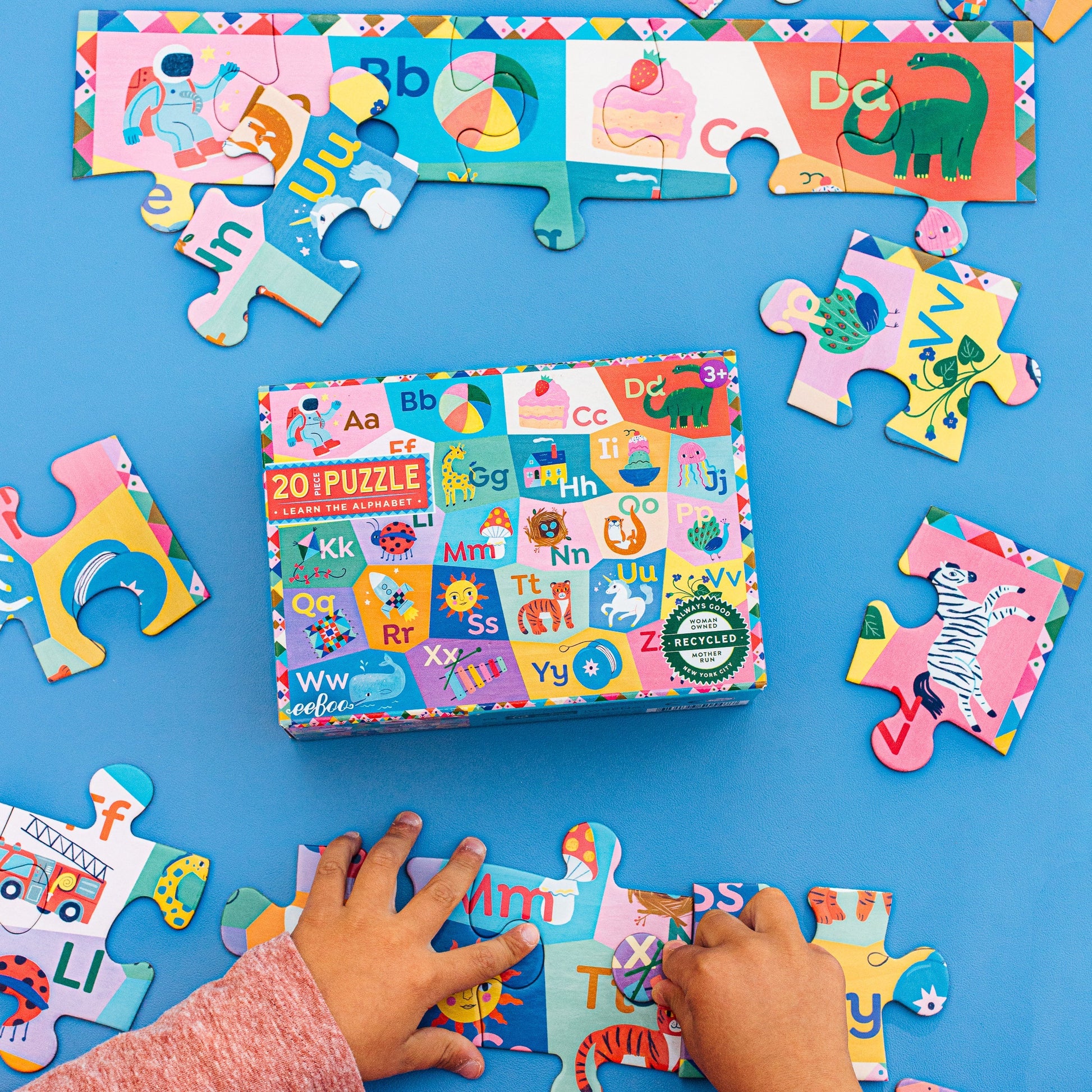 A child assembles Eeboo’s Learn the Alphabet 20 Piece Puzzle with vibrant animal illustrations and letters on a blue surface; completed sections and the puzzle box are also shown.