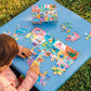 A child assembles the Eeboo Learn the Alphabet 20 Piece Puzzle on a blue outdoor table, with its box and extra pieces nearby.