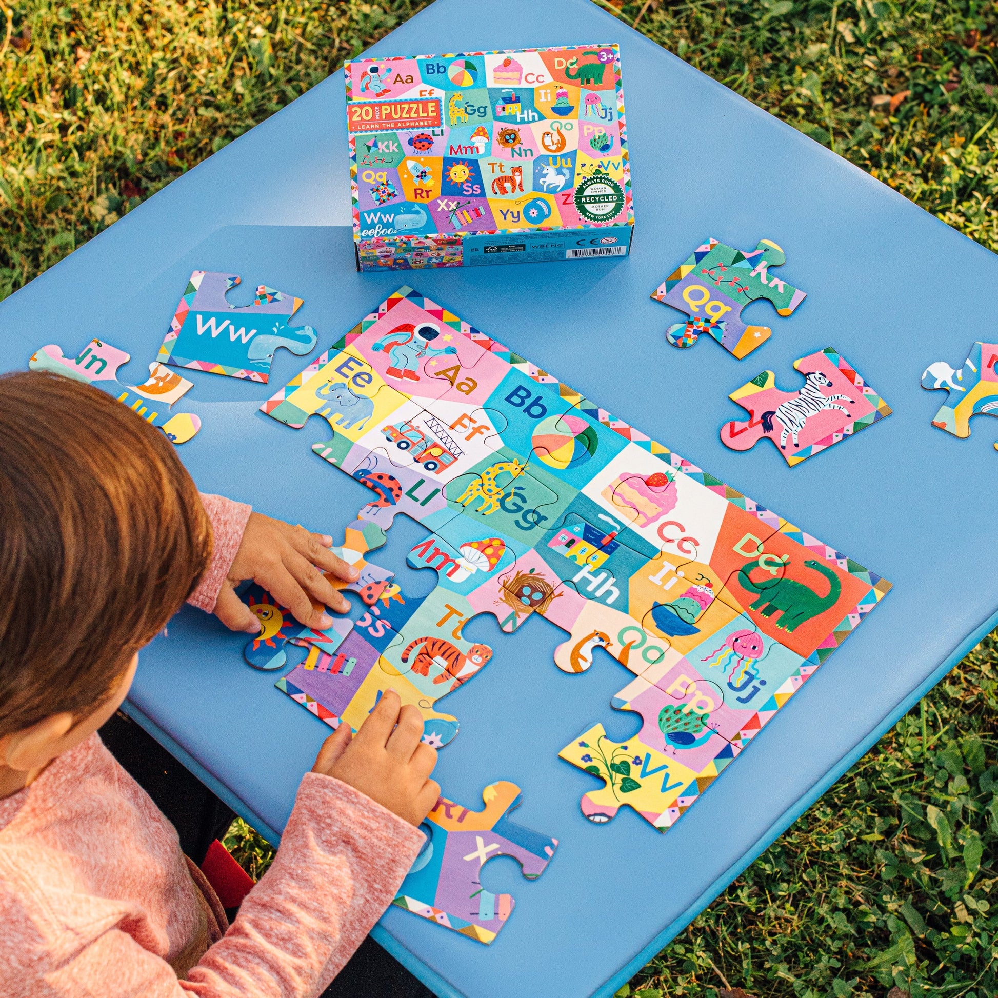A child assembles the Eeboo Learn the Alphabet 20 Piece Puzzle on a blue outdoor table, with its box and extra pieces nearby.