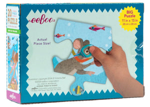 A hand holds a jumbo puzzle piece from the Eeboo Deep Sea Treasure 20 Piece Puzzle, showing a goggle-wearing mouse exploring underwater. The 11 x 15 inch box features a treasure hunt scene for children.