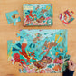 A child's hand assembles pieces from the Eeboo Deep Sea Treasure 20 Piece Puzzle, which showcases a colorful underwater scene with fish and octopuses. This puzzle is designed with jumbo-sized pieces for easy handling, and a box featuring the same vibrant image can be seen in the background.