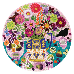 The Eeboo Fruits & Flowers Still Life 500-Piece Round Puzzle showcases a vibrant, floral still life with a vase of flowers, fruit, a bird, and butterflies set against a pink background accented with green, purple, and orange, echoing Vanessa Bowman's distinctive round painting style.