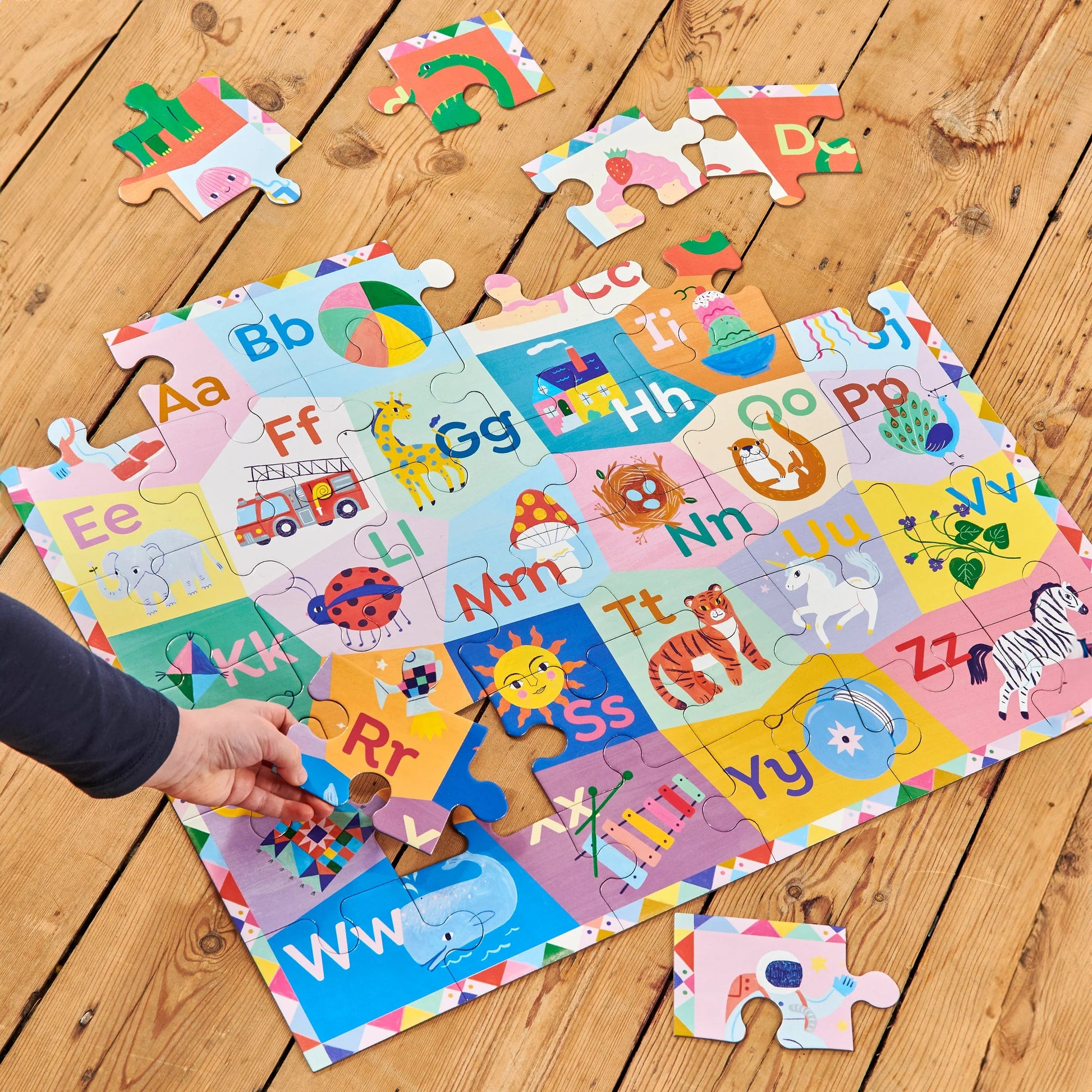 A hand places a piece in the Eeboo Alphabet 36 Piece Giant Puzzle, illustrated by Sarah Walsh and showing animals and objects for each letter, on a wooden floor.