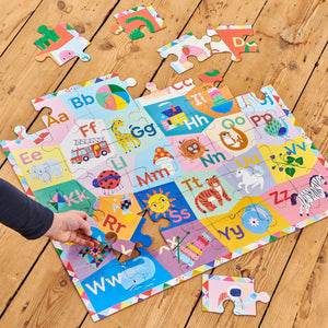 A hand places a piece in the Eeboo Alphabet 36 Piece Giant Puzzle, illustrated by Sarah Walsh and showing animals and objects for each letter, on a wooden floor.