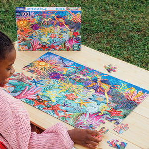 A person assembles the Eeboo Seahorses, Sea Stars & Sea Slugs 100 Piece Puzzle with colorful sea creatures at an outdoor wooden table, as the illustrated puzzle box sits in the background.
