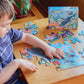 A child eagerly assembles the Eeboo Sharks & Friends 20pc Puzzle on a wooden table, with the Eeboo puzzle box nearby. Among the vibrant pieces, friendly sharks swim into place, capturing preschool attention and sparking curiosity about ocean wonders.