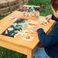 A child joyfully assembles the vibrant eeboo Space Exploration 20 Piece Puzzle on a small wooden table outdoors, with the Eeboo puzzle box nearby.