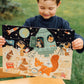 A child proudly holds the completed eeboo Space Exploration 20 Piece Puzzle by Eeboo, featuring a vibrant scene with a rocket and various animals in space suits set against a grassy background. This captivating puzzle combines children's love for animals and puzzles into one delightful experience.