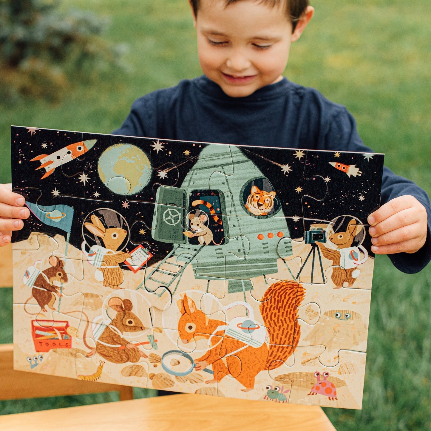 A child proudly holds the completed eeboo Space Exploration 20 Piece Puzzle by Eeboo, featuring a vibrant scene with a rocket and various animals in space suits set against a grassy background. This captivating puzzle combines children's love for animals and puzzles into one delightful experience.