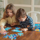 Two children sit at a wooden table assembling the Eeboo Sea Exploration 20-Piece Puzzle, with the puzzle box on the table beside them.