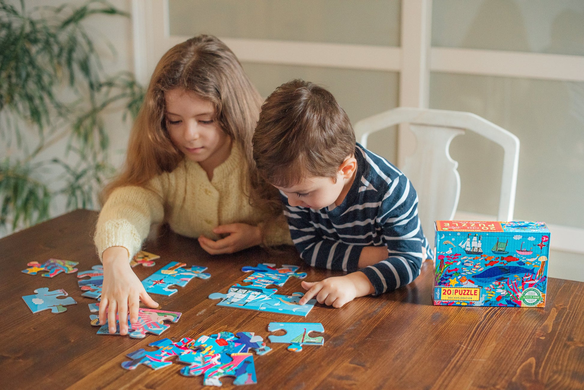 Two children sit at a wooden table assembling the Eeboo Sea Exploration 20-Piece Puzzle, with the puzzle box on the table beside them.