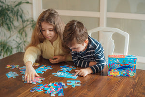 Two children sit at a wooden table assembling the Eeboo Sea Exploration 20-Piece Puzzle, with the puzzle box on the table beside them.