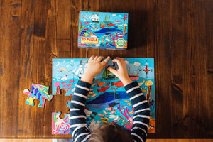A child assembles the Eeboo Sea Exploration 20 Piece Big Puzzle on a wooden table, with the vibrant puzzle box and extra pieces nearby, inspiring curiosity and creativity.