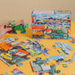 A child's hand holds a puzzle piece from the colorful Eeboo Vehicles 20 Piece Puzzle, which features various vehicles and promotes hand-eye coordination. The puzzle box, displaying an image of the completed city of vehicles, rests on a yellow surface.