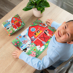 A smiling child sits at a table, putting together the Eeboo Little Farm 20-Piece Puzzle featuring adorable farm animals inspired by Jill Howarth, with the puzzle box and a potted plant also on the table.