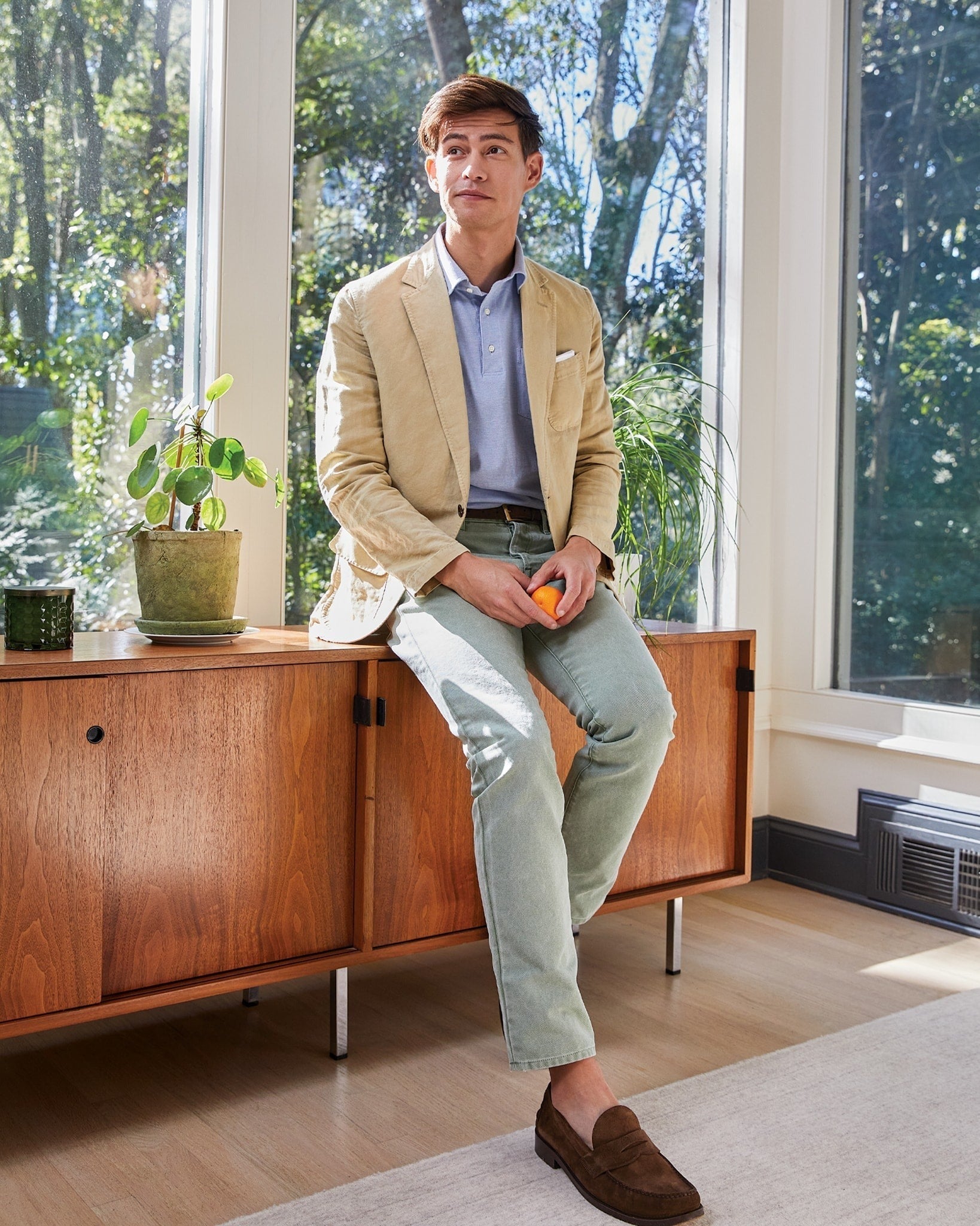 A man in a Sid Mashburn Butcher Jacket, light blue shirt, and light green pants sits on a mid-century modern wooden credenza in a room with large windows. He is holding an orange in his right hand, while his outfit subtly complements the rich texture of Italian canvas.