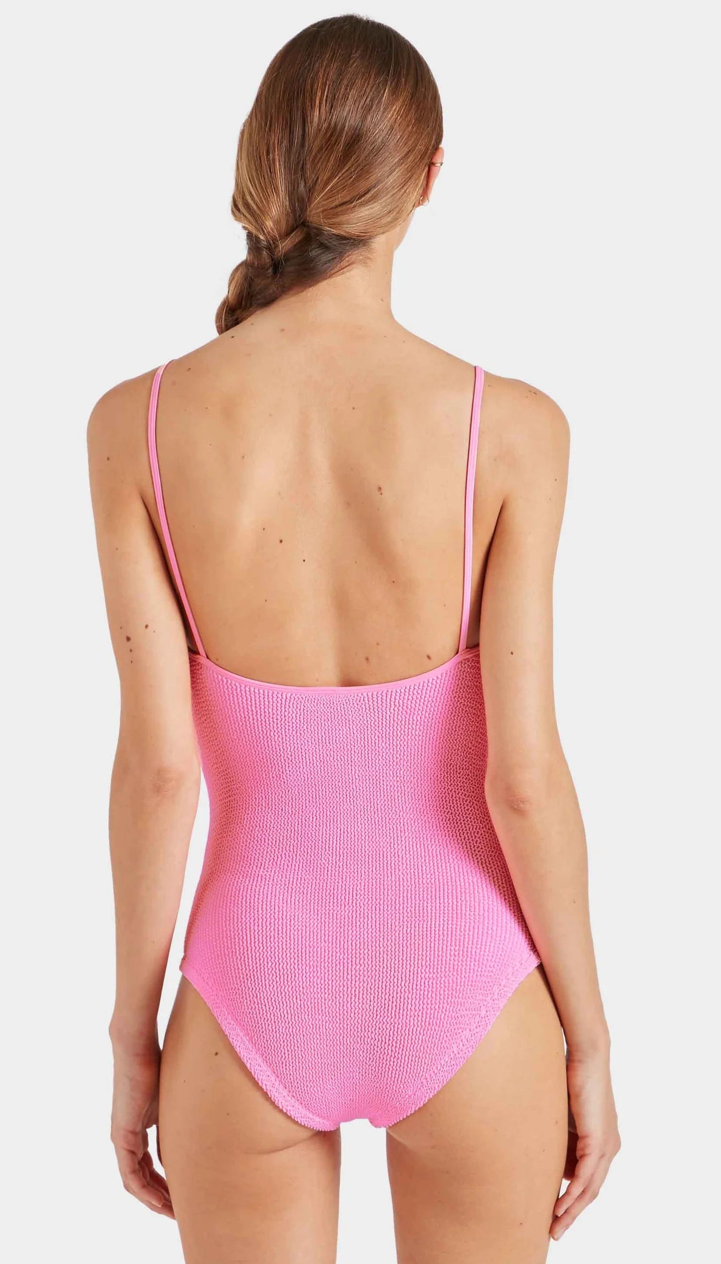 A woman with long brown hair in a braid is shown from the back wearing Hunza G's Pamela Swim, a pink one-piece made from Original Crinkle fabric, with thin shoulder straps and adjustable coverage for a perfect fit.