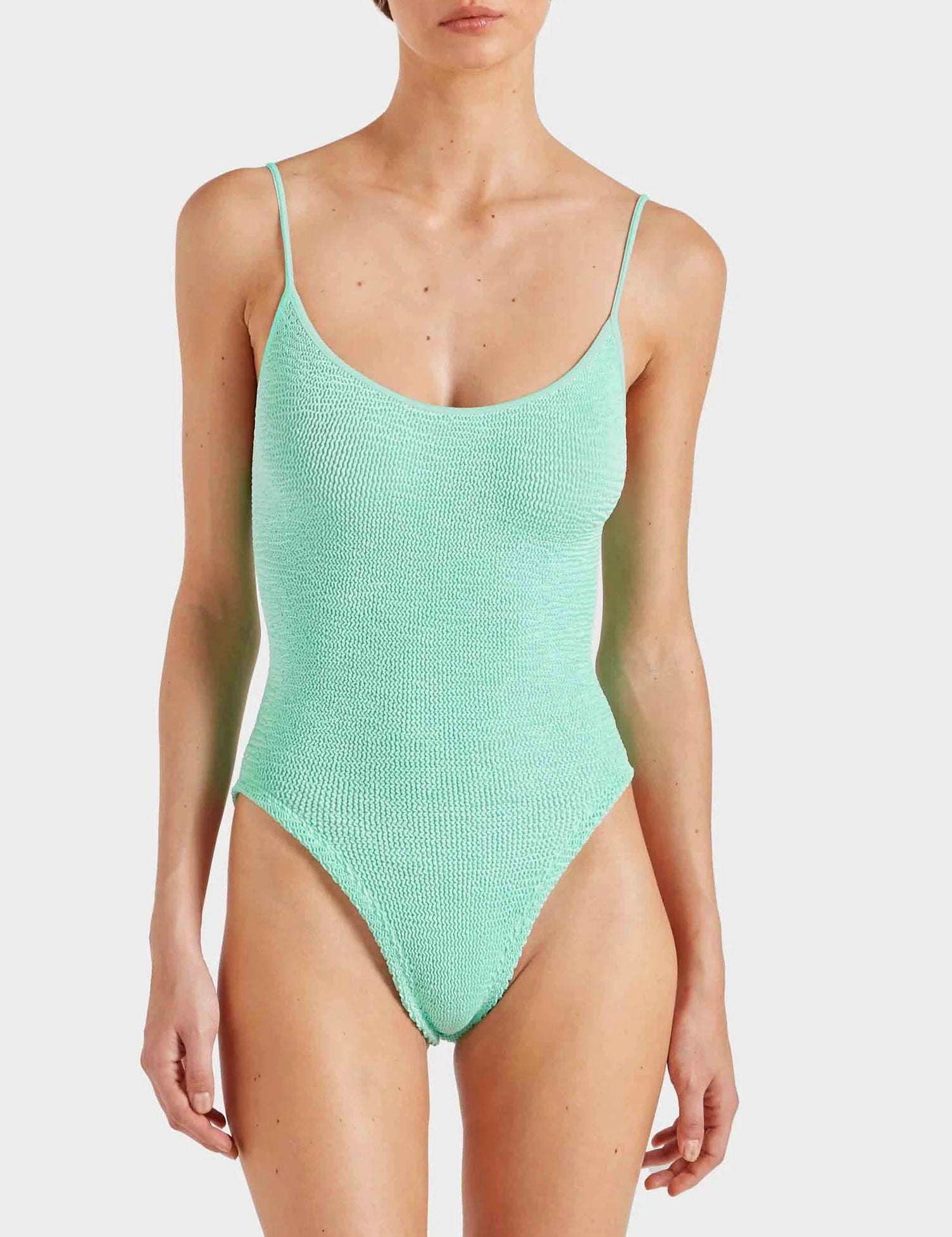 A person is wearing the Hunza G Pamela Swim, a light green one-piece swimsuit made from our signature Original Crinkle™ fabrication with thin straps, standing against a plain background.