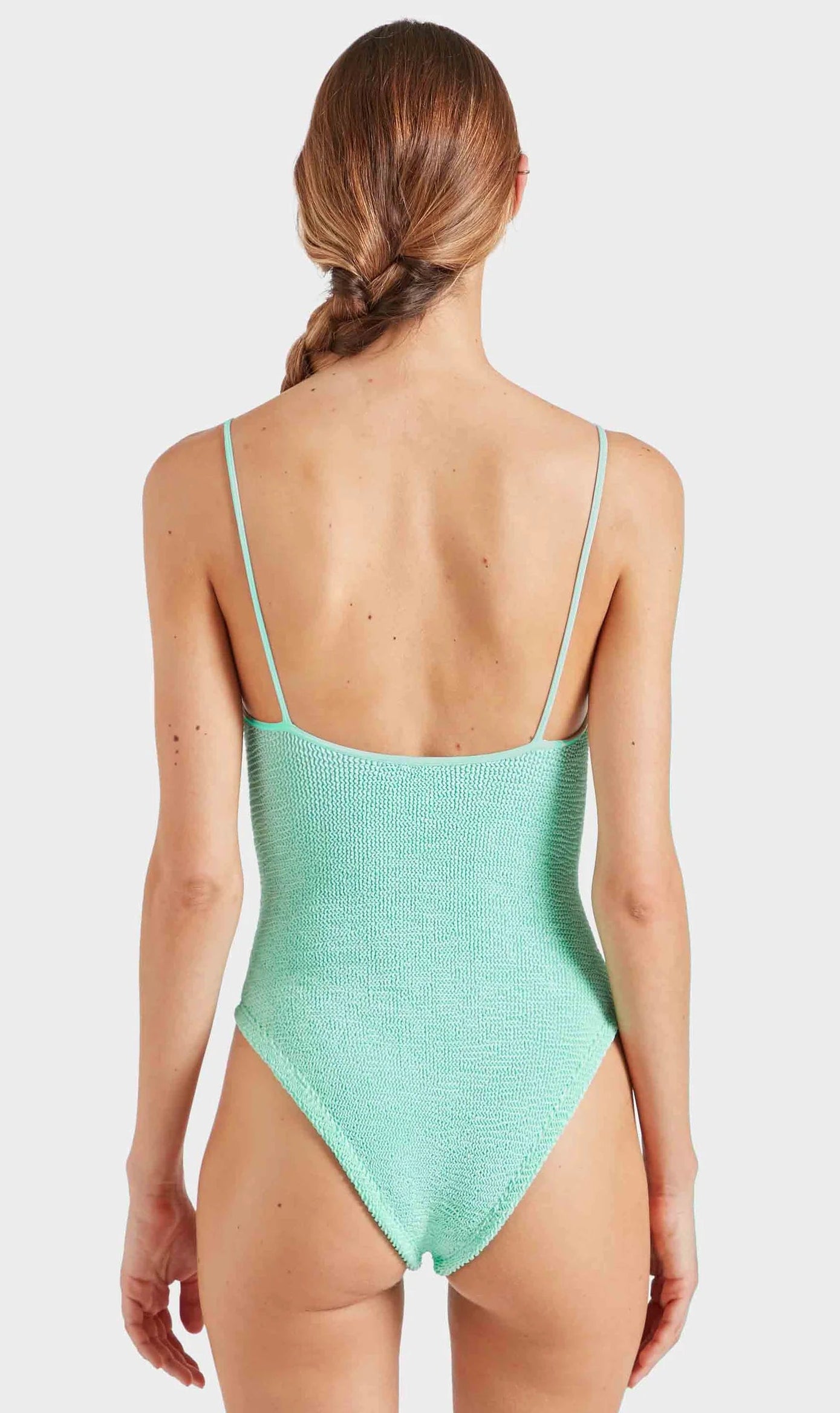 A woman with long brown hair in a braid is shown from the back against a plain white background, wearing the Pamela Swim by Hunza G—a textured light green one-piece swimsuit crafted from Original Crinkle™ fabrication with thin straps.