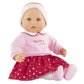 The Corolle Albane Heart Petals Doll, part of the Corolle collection, is crafted from soft vinyl and features a pink headband, a pink top with "Mon Petit" text, and a red skirt decorated with white patterns. The doll is posed against a plain background.