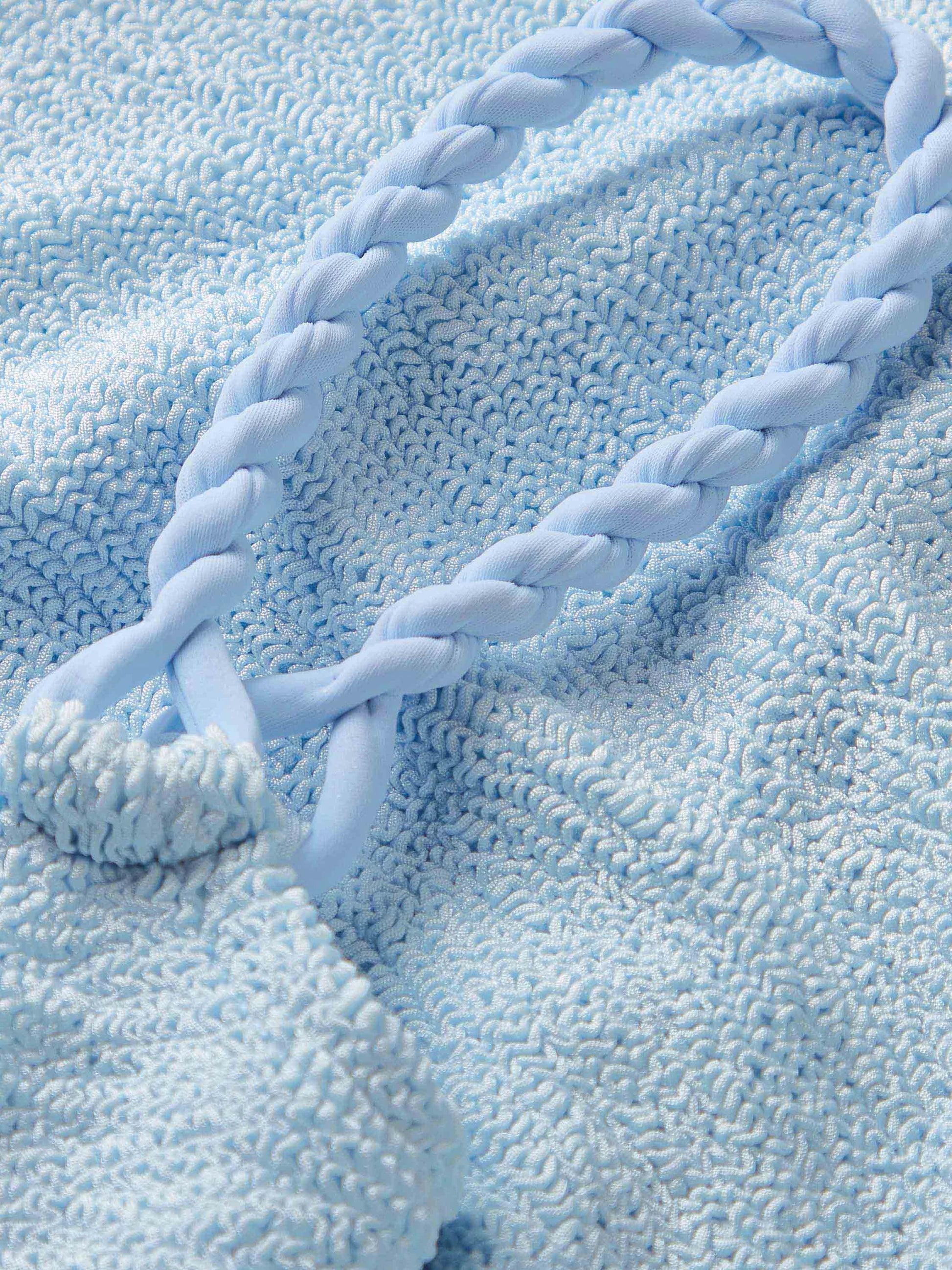 A close-up of the Phoebe Bikini by Hunza G in light blue crinkle fabric, featuring a braided strap, is shown lying on top.