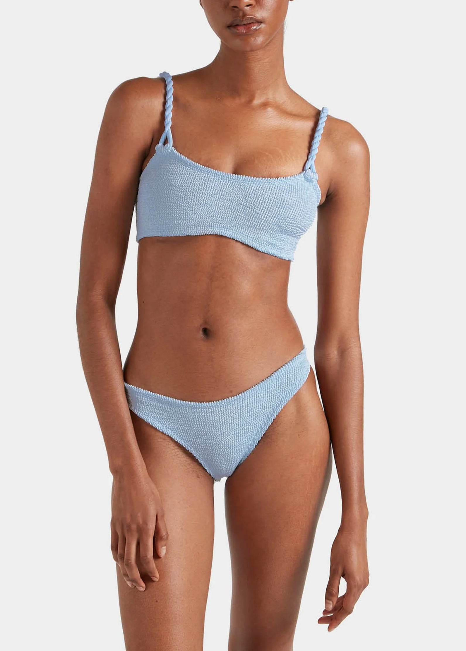 A person wears the Hunza G Phoebe Bikini, featuring textured light blue crinkle fabric and twisted shoulder straps, standing against a plain white background.