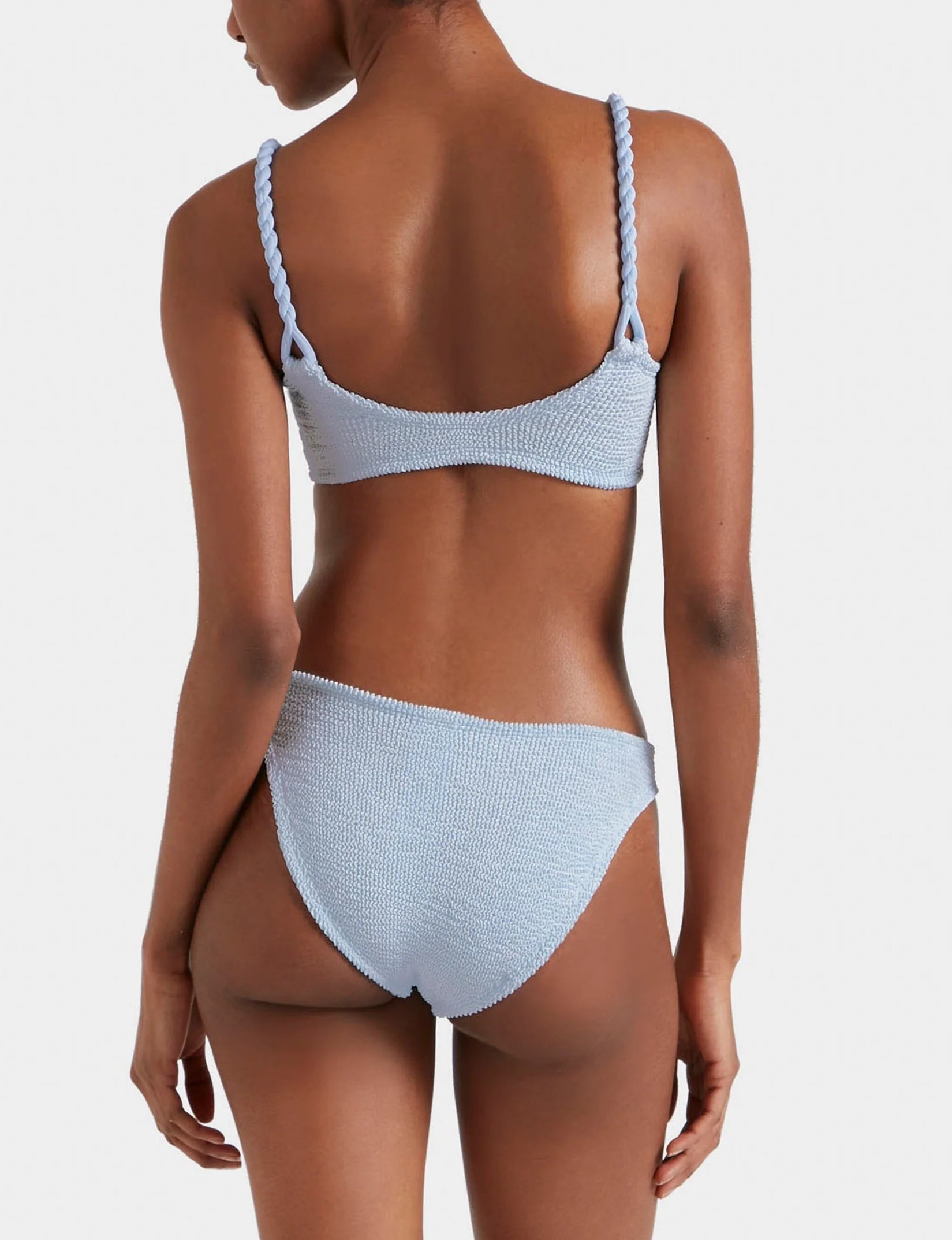 A woman models the Hunza G Phoebe Bikini, featuring light blue crinkle fabric and braided straps, shown from the back against a plain light background.