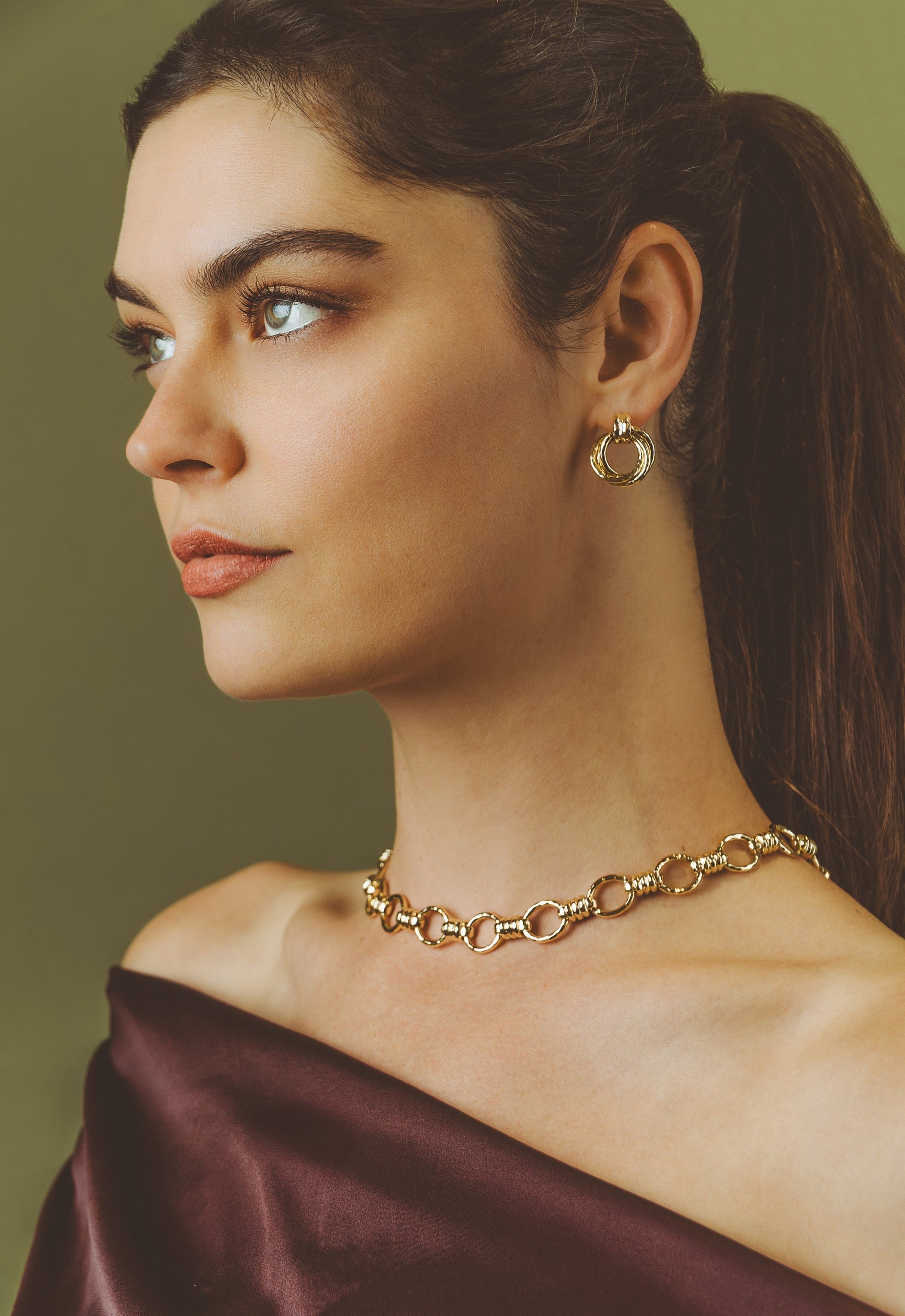 A woman with dark hair in a ponytail, wearing gold hoop earrings and the Anabel Aram Neptune Chain Necklace, pairs it with an off-shoulder burgundy top as she poses against an olive green background.