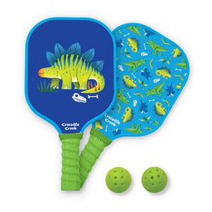 The Crocodile Creek Dinosaur Pickleball Set includes two blue paddle bats with dinosaur designs and green handles, plus two green perforated balls. Perfect for kids ages 6 and up.