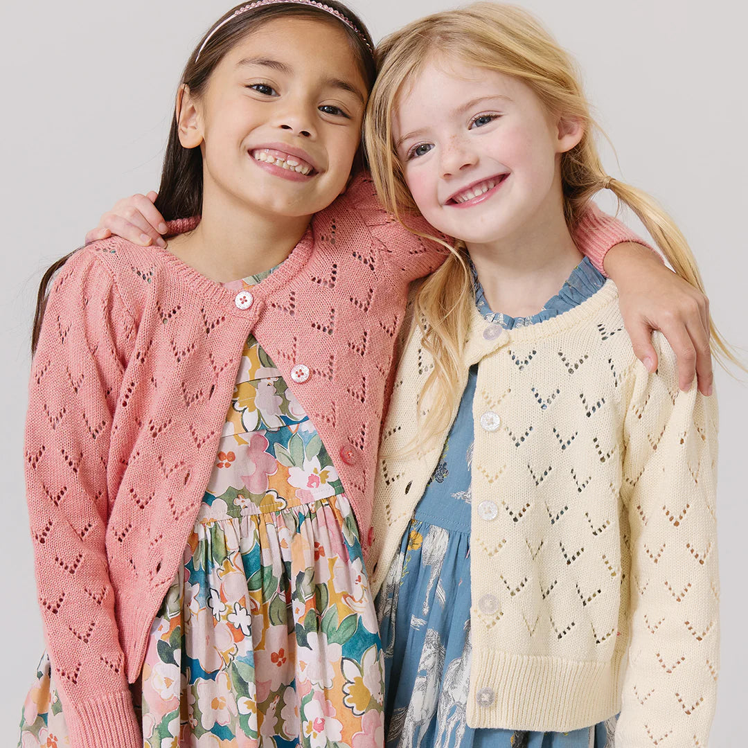 Two young girls stand side by side, smiling with arms around each other. One wears a pink pointelle cardigan and floral dress; the other wears the Pink Chicken Girls' Connie Sweater with glitter buttons over her blue dress.