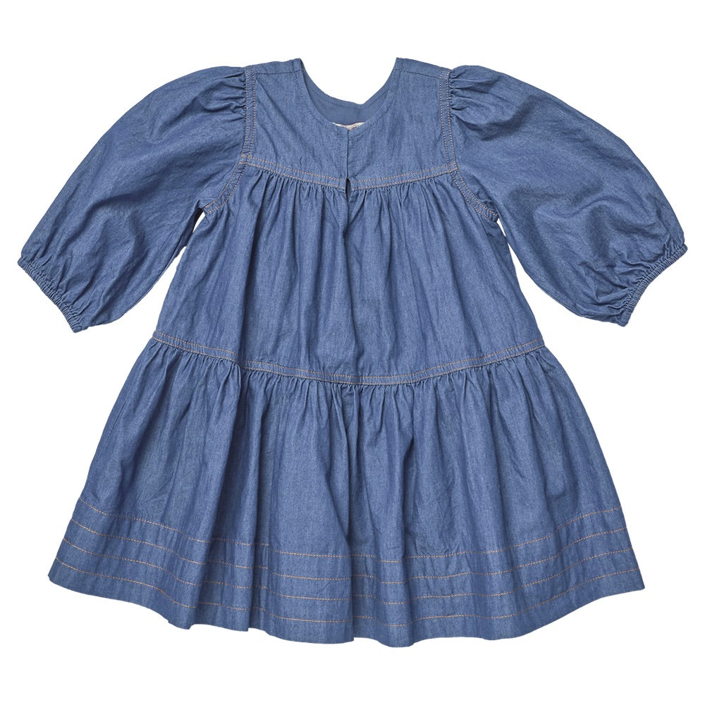 The Pink Chicken Girls’ Sawyer Dress in blue chambray features three-quarter puff sleeves, a gathered waist, and a tiered skirt, displayed on a white background.