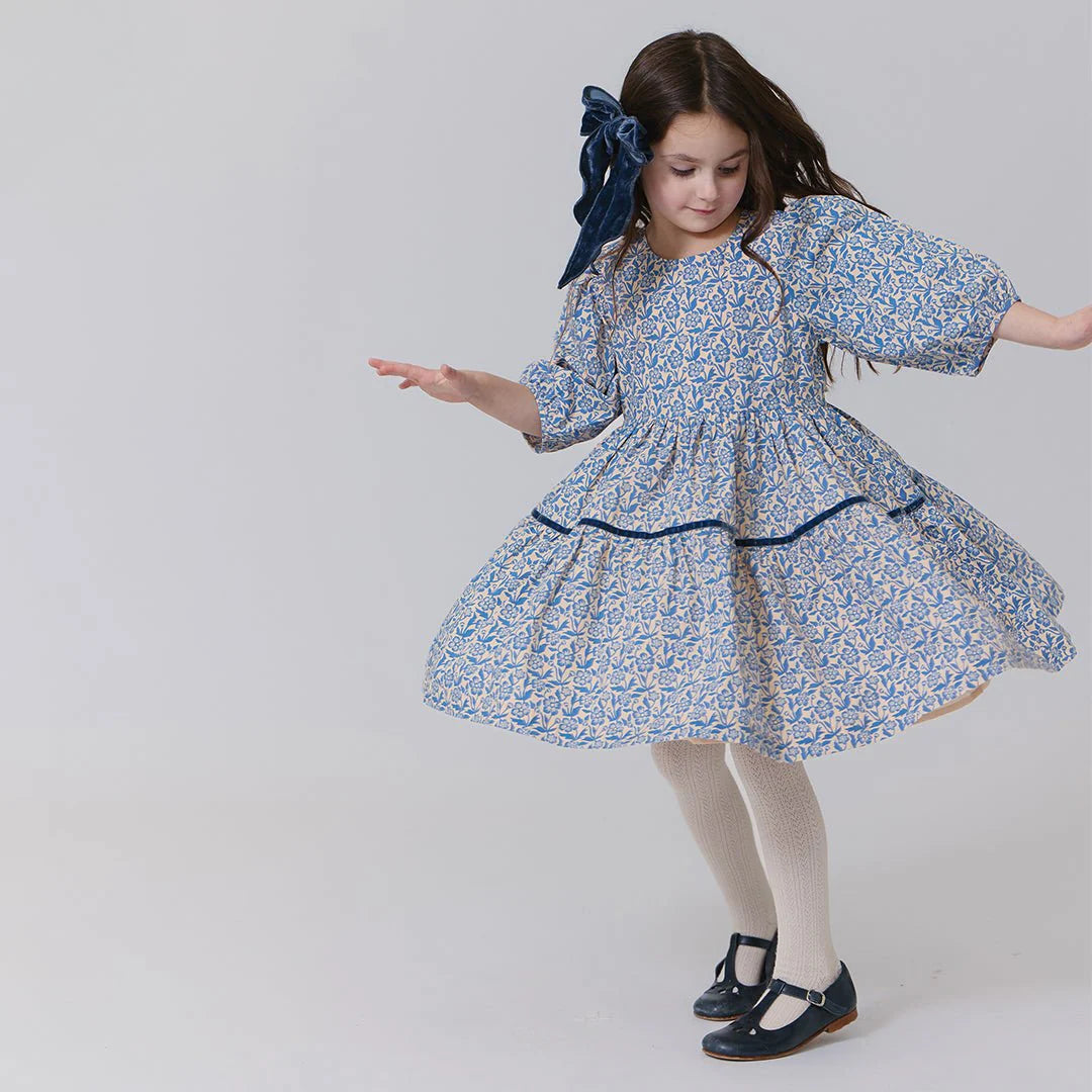 A young girl dances in a Pink Chicken Girls' Maribelle Dress with puff sleeves and climbing floral print, paired with white tights, black shoes, and a large dark blue hair bow against a plain background.