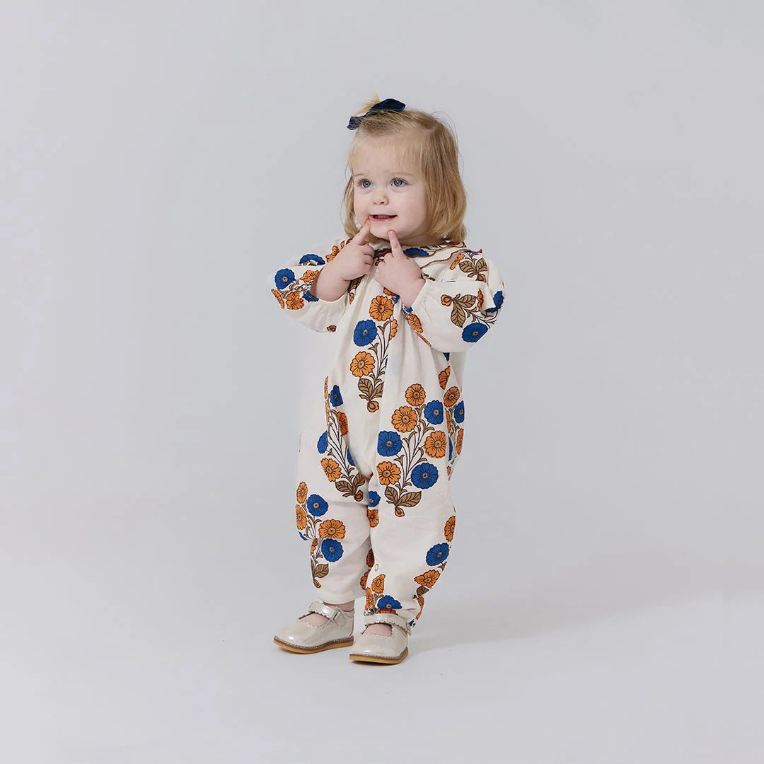 A blonde toddler in a Pink Chicken Baby Girls' Dary Jumper with a scalloped collar and beige shoes stands against a plain light background, holding her fingers to her mouth.