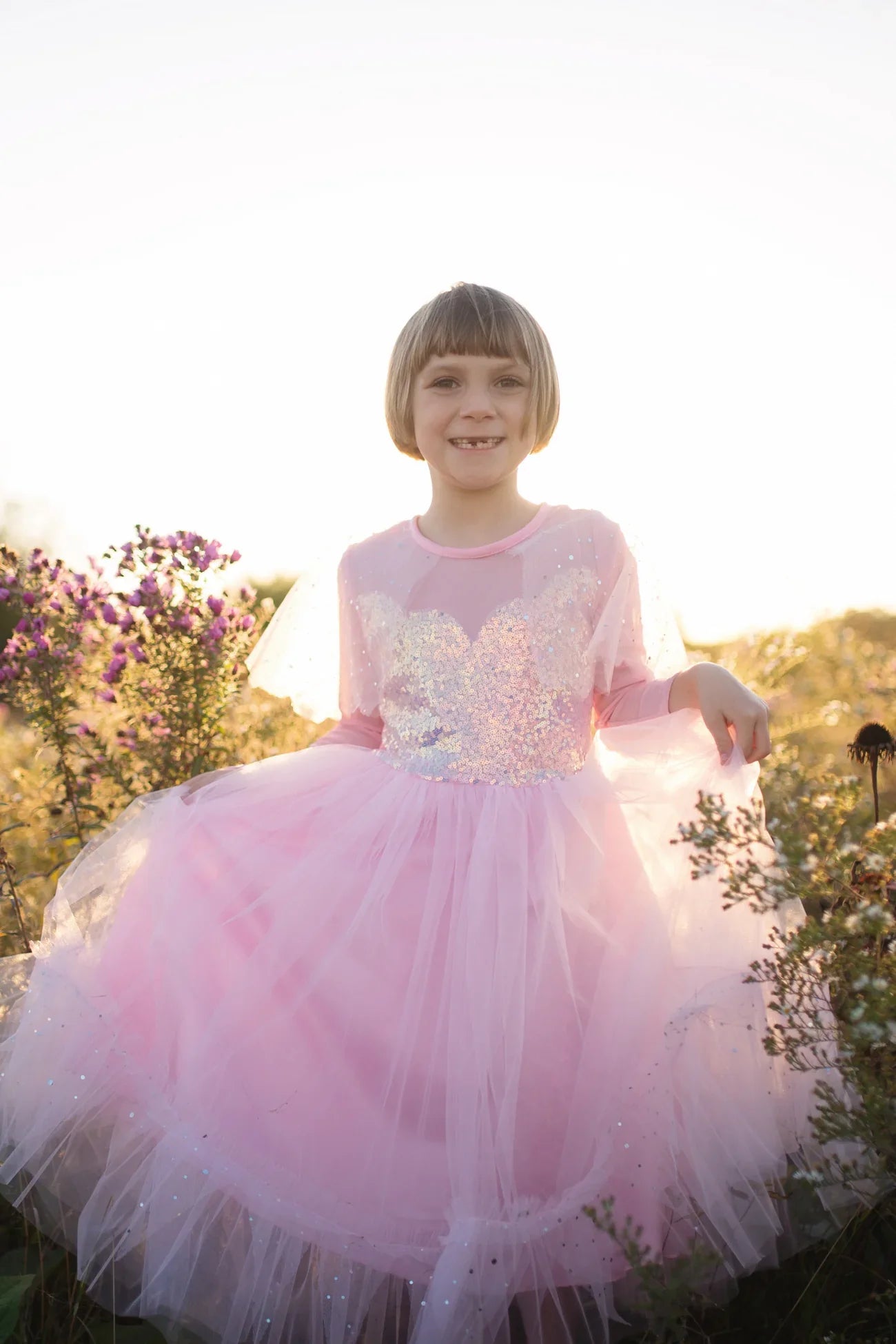 A child wearing the Great Pretenders Elegant In Pink Dress with a sequinned bodice is standing outside in a garden with flowers, smiling at the camera.